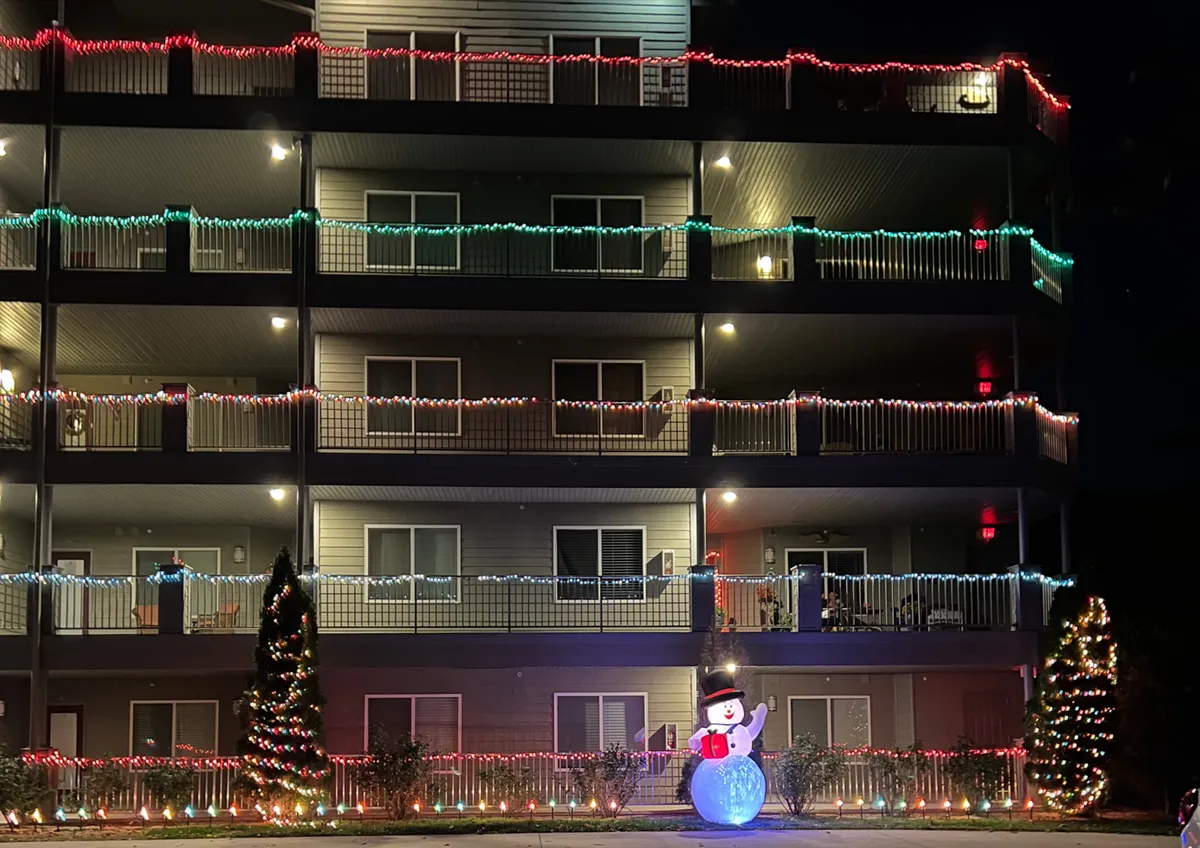 The exterior of the condo building, adorned with enchanting holiday lights at night. A snowman decoration and lit Christmas tree create a welcoming entrance, suggesting a vibrant and community-oriented atmosphere.