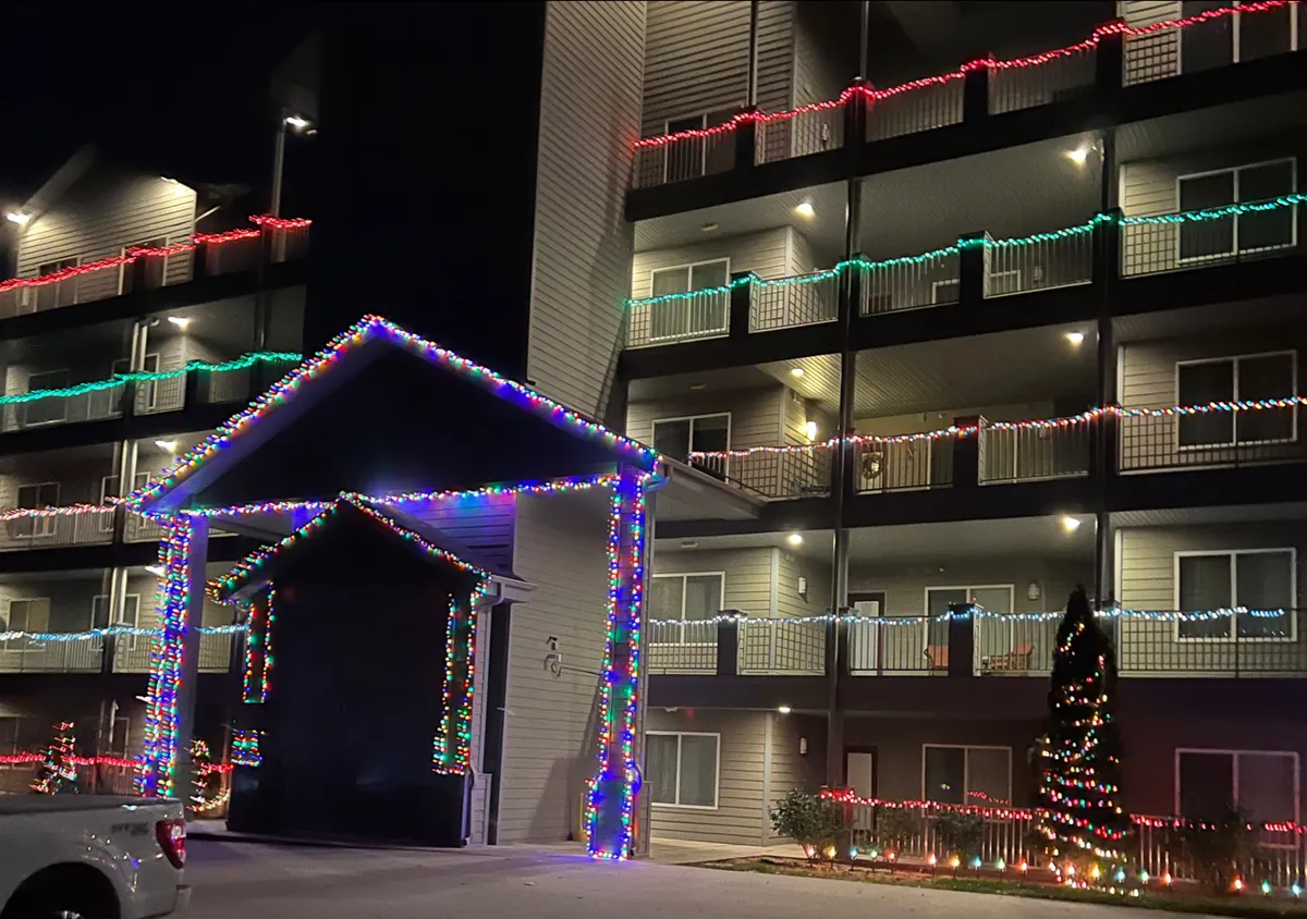 A view of the building’s entrance and balconies, illuminated by colorful holiday lights. The image highlights a decorated exterior and multiple levels, suggesting accessibility via elevators and a vibrant communal environment.
