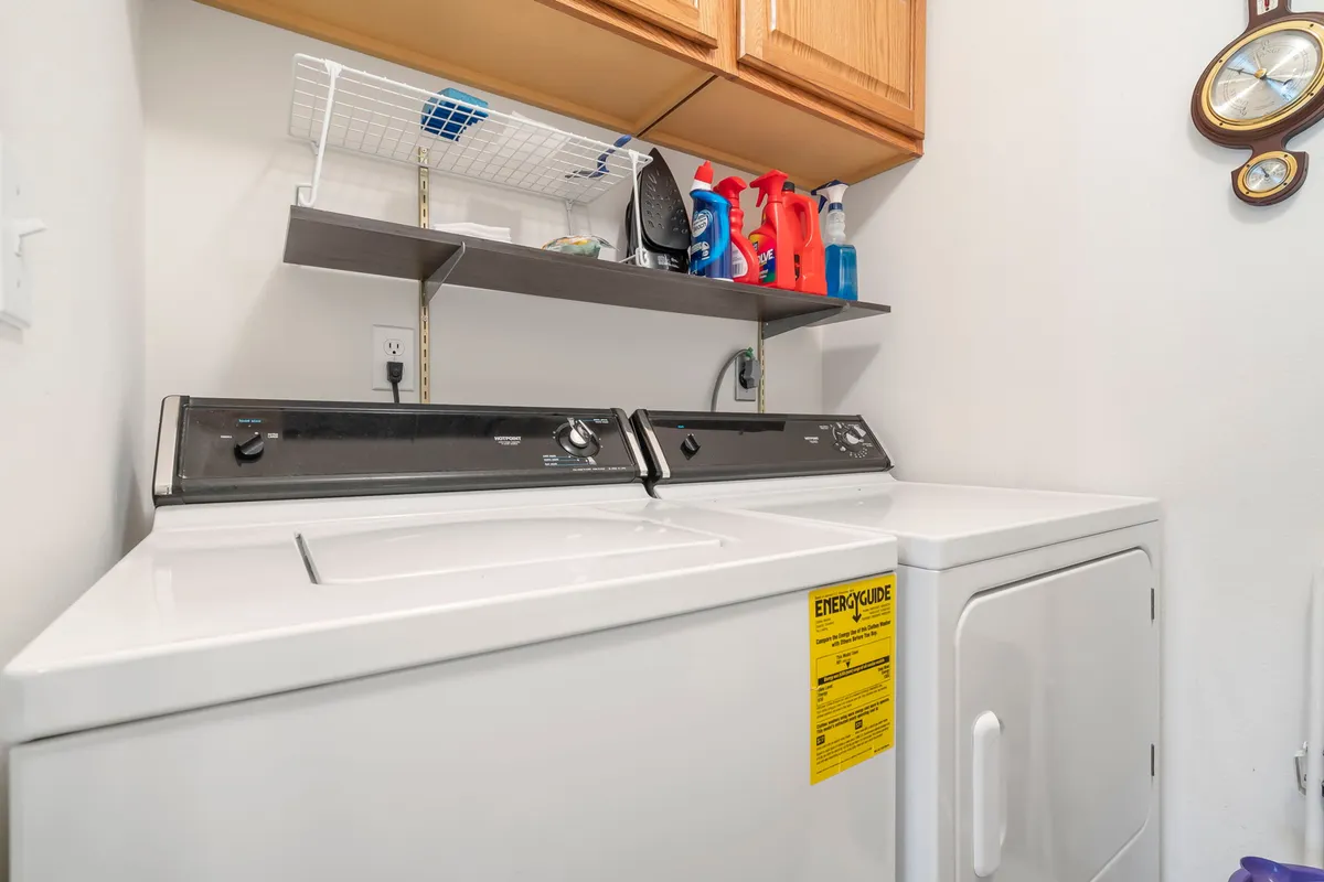 The image displays an in-unit laundry area featuring a washing machine, dryer, shelving unit, and cleaning supplies. The space appears practical and organized, underscoring the convenience of having laundry amenities at home.