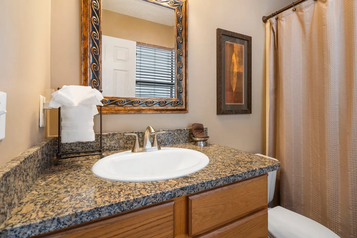 A modern bathroom with a granite countertop, framed mirror, decorative artwork, and a neatly arranged towel holder. The clean and inviting design provides a sense of spa-like relaxation and comfort.