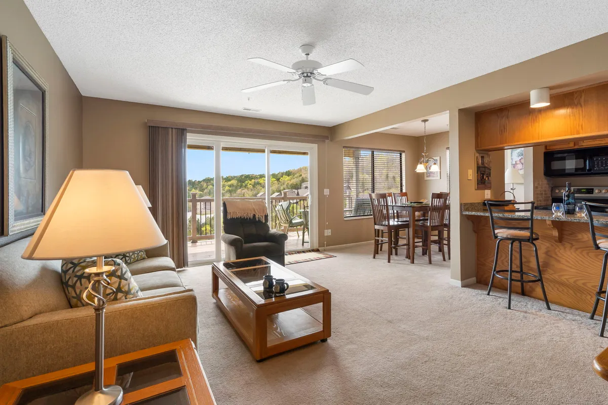 A cozy, well-appointed living space featuring a sofa, dining table, and window view of the deck and lake beyond. The intimate setup is designed for socializing, dining, and enjoying sunsets, with warm natural light enhancing the atmosphere.
