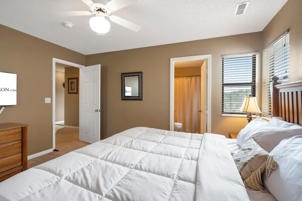 A spacious king bedroom featuring a bed with crisp white bedding, a nightstand with a lamp for ambient lighting, a TV situated atop a dresser, a wall mirror, and doorways leading to other rooms. The room exudes comfort and offers plenty of space for relaxing or enjoying personal downtime.