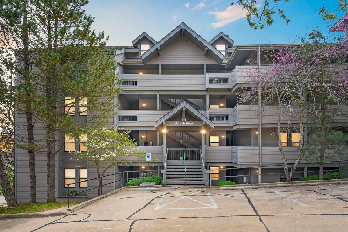 The photo features the charming property entrance with a pitched roof, surrounded by mature trees and a welcoming parking area. The image conveys a sense of arrival and comfort, suggesting easy returns between outings and a cozy atmosphere to come home to.
